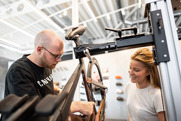Ein Fahrradmechaniker arbeitet an einem Rennrad. Neben ihm steht eine Frau mit weissem T-Shirt.